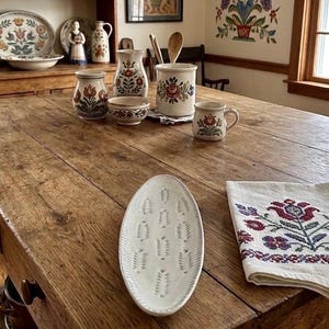 May include: A rustic wooden table displays a collection of vintage ceramic tableware. The set includes a serving platter, small bowls, mugs, and vases, all adorned with floral patterns in red, blue, and green. A floral embroidered cloth adds a touch of color.
