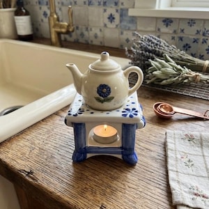 May include: A small, cream-colored ceramic teapot with a blue floral design sits atop a blue and white ceramic warmer. A lit tea light candle is inside the warmer. The scene is set on a wooden surface with a floral patterned napkin.
