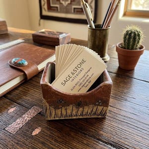 May include: A handmade, rectangular ceramic business card holder with a brown and tan color scheme. The holder contains business cards with the text "SAGE & STONE INTERIORS". A small cactus in a terracotta pot sits nearby on a wooden desk.