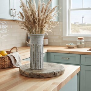 May include: A tall, cylindrical, light gray ceramic vase with vertical fluted ridges and a flared rim, filled with a large arrangement of dried beige grasses and wheat stalks, resting on a circular, natural-edged wooden slab base.