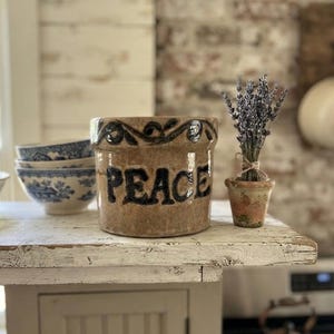 May include: A cylindrical tan ceramic crock featuring the word PEACE in bold black lettering on the front, with a decorative black scroll pattern around the top rim. The piece has a glossy finish and a straight-sided design.