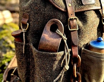 May include: A brown tweed backpack with leather detailing and straps. The backpack features multiple pockets, a wooden chain, and a wooden handle. The label reads "Wilderness Craft". A blue and brown water bottle is in the side pocket.
