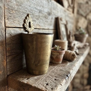 May include: A brass wall-mounted vessel featuring a tapered cylindrical body and a decorative fan-shaped mounting plate at the top. The metal surface displays a weathered, aged patina with subtle horizontal lines etched around the upper rim.
