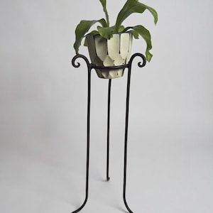 May include: A tall, dark brown metal plant stand with three slender legs and curled top accents. The stand holds a cream-colored ceramic pot with a geometric, faceted exterior pattern and dark vertical lines, containing a green leafy plant.
