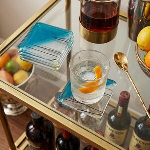 May include: A bar cart with a gold frame and glass shelves. It holds a stack of square coasters with a blue and white design, a glass of water with an orange peel, and bottles of liquor. A gold spoon and a bowl of fruit are also visible.