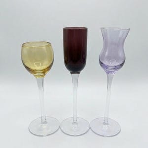 May include: Three colorful stemmed glasses. The glass on the left is yellow, the middle glass is dark red, and the glass on the right is purple. All three glasses have clear stems and bases, and are set against a white background.
