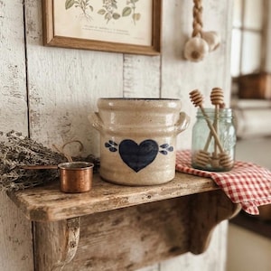 May include: A beige ceramic crock with two side handles and a dark blue heart design on the front. The crock is positioned on a wooden shelf alongside a small copper measuring cup, a glass jar containing wooden honey dippers, and a red checkered cloth.