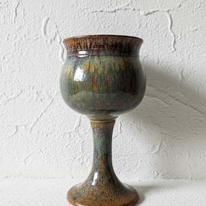 May include: A handmade ceramic goblet with a wide bowl and a slender stem. The goblet features a blend of earthy tones, including brown, green, and blue, with a glossy finish. The base is approximately 3 inches in diameter.