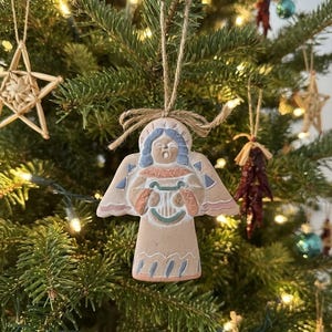 May include: A flat, tan-colored clay angel ornament featuring blue hair, a white harp, and pink and blue geometric accents. The angel is suspended by a natural jute twine string tied in a bow at the top, with a carved design on the front surface.