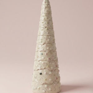 May include: A cream-colored, ceramic decorative tree with a conical shape. The tree is textured with small, raised dots and layered sections. It has a speckled appearance with subtle hints of color, set against a soft pink background.