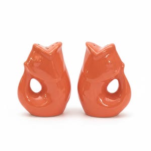 May include: A pair of orange ceramic salt and pepper shakers. Each shaker has a unique, organic shape with a large, circular opening and a pointed top with three small holes. The shakers are a vibrant, solid orange color.