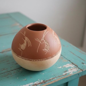 May include: A round, terracotta-colored ceramic vase with a cream-colored base. The vase features etched designs of figures with long limbs. The vase sits on a distressed, turquoise-painted wooden table.