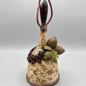 May include: A decorative bell with a brown wooden handle and a burgundy rope. The bell is adorned with a brown and tan textured pattern, a wreath of brown accents, and a cluster of nuts and green leaves. The base has a woven brown trim.