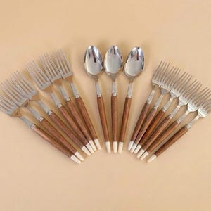 May include: A set of stainless steel forks and spoons with wooden handles and white tips. The silverware is arranged in a fan shape against a neutral background. The spoons have a rounded bowl shape.