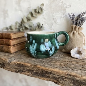May include: A rounded ceramic mug featuring a glossy deep green glaze with white crystalline patterns. The mug has a wide body, a smooth interior, and a sturdy curved handle on the right side, showcasing a distinct reactive finish on its surface.