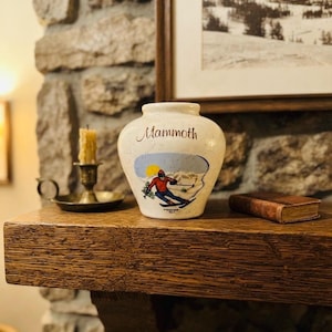 May include: A cream-colored ceramic vase with a speckled finish, featuring the word Mammoth in script and a graphic of a skier in a red jacket and blue pants on a snowy slope under a yellow sun. The vase has a rounded body and a short neck.