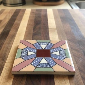 May include: Square ceramic tile with a geometric design. The tile features a central dark red square surrounded by a pattern of triangles, rectangles, and curved shapes in shades of pink, blue, green, and cream. The design includes white polka dots and floral motifs.