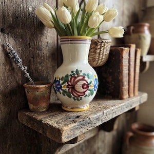 May include: A cream-colored ceramic vase with a hand-painted floral motif in red, green, and blue, containing a bouquet of white tulips. The vase features a flared rim and a rounded body, resting on a wooden shelf alongside a small terracotta pot.