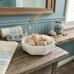 May include: A white, octagonal bowl filled with various seashells sits on a weathered wooden table. A blue and white striped towel and a bar of soap are also visible. The scene evokes a coastal, beach-themed aesthetic.