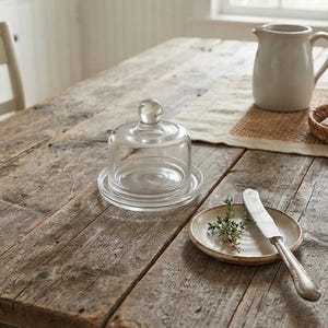 May include: A clear glass butter dish with a matching dome lid featuring a spherical knob handle. The dish sits on a flat base with a raised rim, designed to hold a stick of butter. The glass is transparent, smooth, and circular in shape.