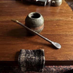 May include: A round, metallic mate gourd featuring an embossed floral pattern and a copper-colored band around the center. A long, slender metal bombilla straw with a gold-toned ring and a perforated, spoon-shaped filter end rests across the gourd.