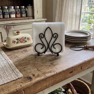 May include: A black metal napkin holder with a decorative scrollwork design, featuring a symmetrical pattern of curved lines and small ball feet, holding a stack of plain white square paper napkins in an upright position.