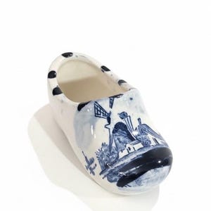 May include: A small, white ceramic clog with a blue and white windmill design. The top edge has black polka dots. The Dutch-style shoe is a decorative item, likely a souvenir or collectible.