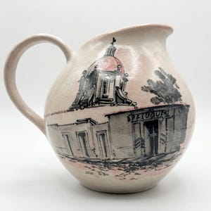 May include: Cream-colored ceramic pitcher with a rounded body and a handle. The pitcher features a hand-drawn illustration of a building with a red-domed roof and the word "SPETUOFK" above the entrance. The design is in black and gray.