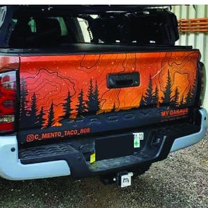 May include: A truck bed with an orange and black mountain and forest design. The design includes the text "MY GARAGE" and an Instagram handle. The truck has large tires and a black bumper.
