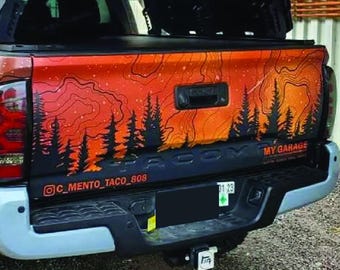 May include: A truck bed with an orange and black mountain and forest design. The design includes the text "MY GARAGE" and an Instagram handle. The truck has large tires and a black bumper.