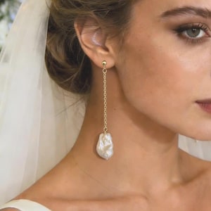 May include: A gold chain earring with a large, irregular white pearl pendant. The earring is worn by a person with light skin, brown hair, and a white dress. The background includes white flowers and a chandelier.