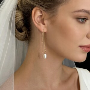 May include: A woman wearing gold drop earrings with a pearl at the end of each chain. The earrings are delicate and elegant, with a simple design. The woman is wearing a white dress and a veil, suggesting a bridal theme.