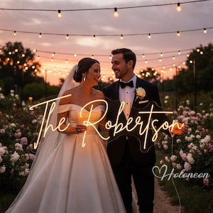 Puede incluir: Un letrero de neón personalizado con el texto The Robertson en una tipografía cursiva de color amarillo cálido, montado sobre una base de acrílico transparente. El letrero es sostenido por una pareja vestida de boda en un jardín al atardecer.