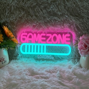 Może przedstawiać: Neonowy znak z napisem "GAME ZONE" w kolorze różowym nad niebieskim paskiem ładowania. Słowo "LOADING" znajduje się poniżej. Znak jest na puszystej białej powierzchni, z kwiatami w wazonach.