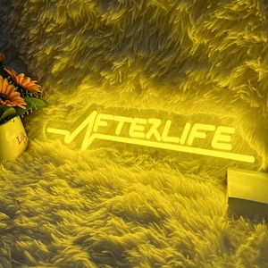 Puede incluir: Un letrero de neón amarillo con la palabra "AFTERLIFE". El letrero presenta un diseño de línea de latido cardíaco sobre el texto. Se coloca sobre un fondo amarillo y esponjoso.