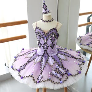 Puede incluir: Un tutú de ballet en tonos lavanda y blanco, adornado con ribetes de encaje negro, adornos brillantes y detalles de cuentas. El disfraz incluye un tocado y gargantilla a juego, exhibidos en un maniquí. El corpiño presenta detalles en forma de diamante.