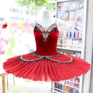 Może przedstawiać: Czerwone baletowe tutu z aksamitnym stanikiem i warstwową, błyszczącą spódnicą. Stanik jest ozdobiony wyszukanymi koralikami i kryształami. Tutu jest prezentowane na manekinie, prezentując jego design i konstrukcję.