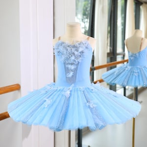 May include: A pale blue ballet tutu with a fitted bodice and a layered skirt. The bodice is decorated with floral details and the skirt has glitter accents. The tutu is displayed on a mannequin, with a similar tutu visible in the background.