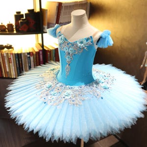 Może przedstawiać: Jasnoniebieskie baletowe tutu i gorset wystawione na manekinie. Gorset jest ozdobiony kwiatowymi i srebrnymi zdobieniami. Tutu ma wiele warstw tiulu, z kwiatowymi i kryształowymi akcentami. Kostium jest przeznaczony do występów tanecznych.