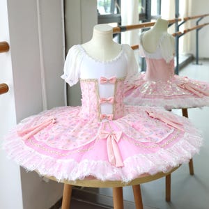 May include: A pink and white ballet tutu with a bodice adorned with bows and gold trim. The tutu features layers of pink fabric, lace trim, and decorative embellishments. A second tutu is visible in the background.