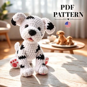 May include: A crocheted Dalmatian dog with black spots on a white body. The dog has a black nose, pink paws, and a smiling expression. The image includes the text "PDF PATTERN" and a small American flag.