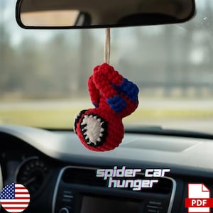 Op de afbeelding: Een rode, blauwe en witte gehaakte auto-hanger in de vorm van een spin, hangend aan een achteruitkijkspiegel. De tekst "spider car hunger" is zichtbaar op het dashboard.