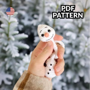 Könnte beinhalten: Ein kleiner, weiß gehäkelter Schneemann mit Karottennase, schwarzen Augen und einem lächelnden Mund. Der Schneemann hat drei schwarze Knöpfe und wird in einer Hand gehalten. Der Text "PDF PATTERN" befindet sich oben rechts.