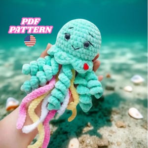 May include: A crocheted teal jellyfish with a smiling face and colourful tentacles. The jellyfish has a red heart-shaped detail and is labelled "PDF PATTERN" with an American flag icon.