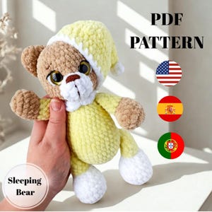 May include: A crocheted teddy bear wearing a yellow nightcap and pajamas. The bear has brown fur on its head and limbs, with white accents on the paws and face. The text "PDF PATTERN" and "Sleeping Bear" are visible.