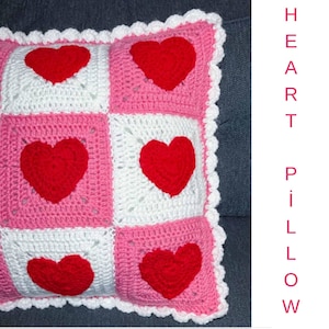 May include: A handmade crochet pillow with a patchwork design. The pillow features alternating squares of pink and white, each with a red heart in the center. The edges are trimmed with a white scalloped border. The text "HEART PILLOW" is visible.