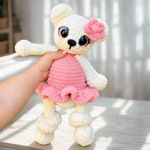 May include: A handmade white crochet bear doll with a pink dress and flower. The bear has large black eyes, a black nose, and a pink flower on its head. The doll is held in a person's hand.