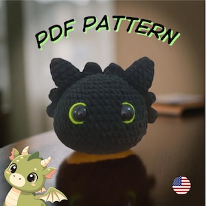 May include: A black crocheted dragon head with large, round eyes outlined in neon green. The text "PDF PATTERN" is above the dragon. A cartoon dragon is in the lower left corner. The dragon head is a handmade item.