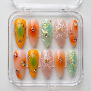 May include: A set of ten artificial nails in a clear plastic case. The nails feature various designs, including gradients of orange, yellow, and green, with silver and gold accents. Some nails have floral or swirl patterns, and all are oval-shaped.