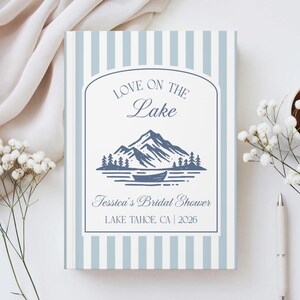 Pode incluir: Uma placa retangular com listras verticais azul-claras e brancas com um design de arco central. O texto diz LOVE ON THE Lake, Tessica's Bridal Shower e LAKE TAHOE, CA | 2026, com um gráfico azul de montanhas e uma canoa.